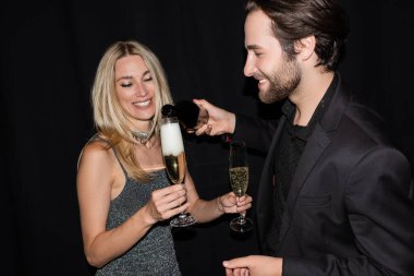 Smiling man in jacket pouring champagne near blonde girlfriend during party isolated on black 