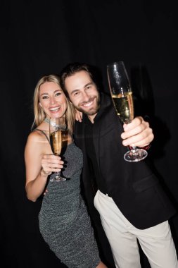 Positive couple looking at camera while holding glasses of champagne isolated on black 