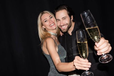 Blonde woman in dress hugging boyfriend and holding glass of champagne isolated on black 