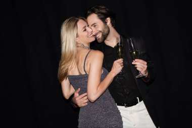 Smiling blonde woman holding glass of champagne and hugging stylish boyfriend isolated on black 