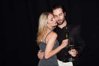 Sexy woman in dress holding glass of champagne and hugging boyfriend isolated on black 