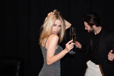 Sexy woman in dress holding champagne while dancing near boyfriend isolated on black 