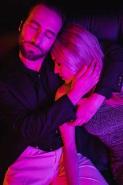 Young man hugging blonde girlfriend on leather couch with lighting isolated on black 