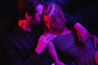 High angle view of sexy man touching blonde girlfriend on leather couch with lighting isolated on black 