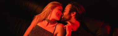 side view of sexy woman kissing bearded boyfriend on leather couch with lighting isolated on black, banner 