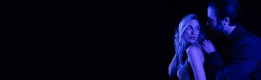 Young bearded man touching blonde girlfriend with blue lighting isolated on black, banner 