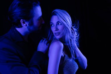Sexy blonde woman looking at boyfriend with blue lighting isolated on black 