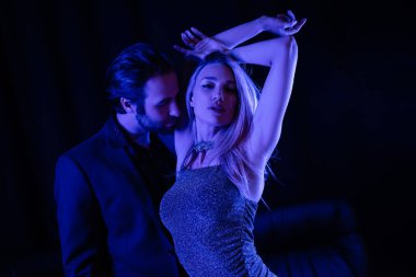 Passionate man standing near sensual girlfriend in dress with blue lighting on black background 