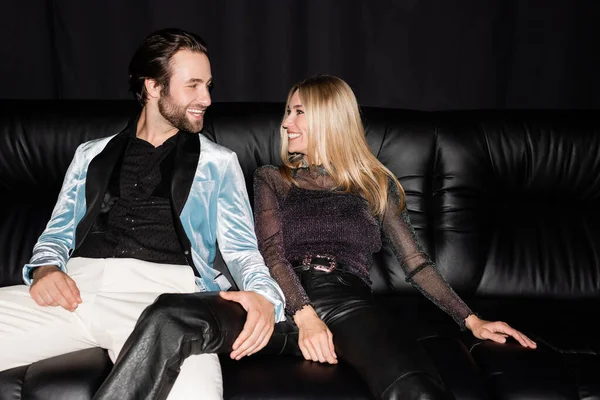 Cheerful stylish couple looking at each other while sitting on leather couch isolated on black 