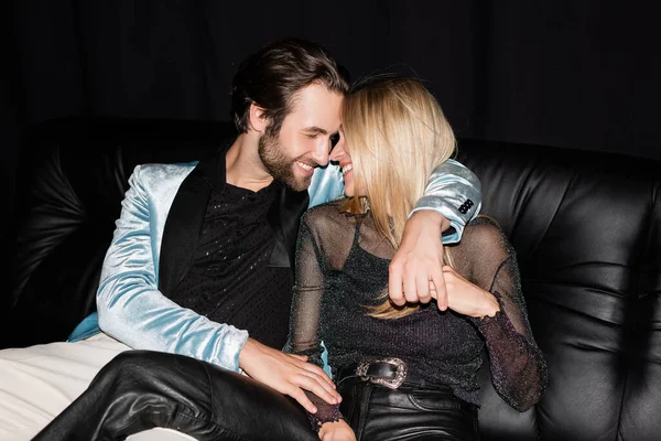 Positive man in jacket hugging blonde girlfriend on leather couch isolated on black 