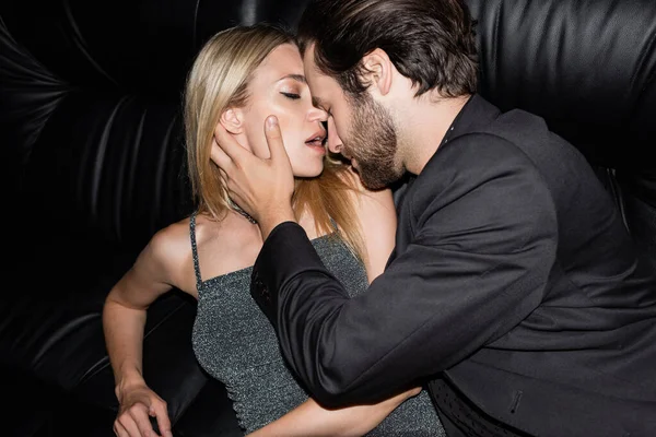 Bearded man in jacket touching sensual blonde girlfriend on leather couch