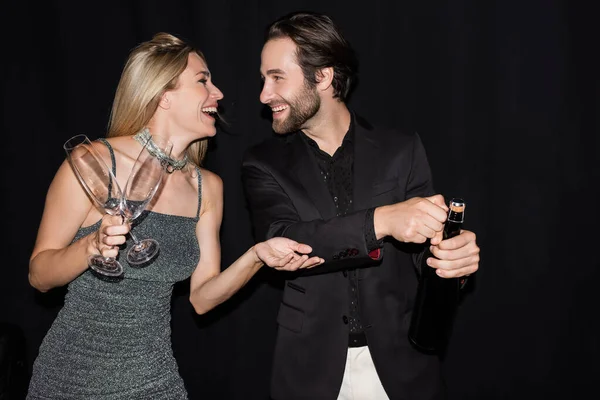 Side view of positive blonde woman in dress holding glasses near boyfriend with champagne isolated on black