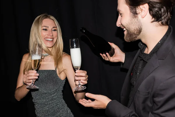 Smiling blonde woman holding glasses while boyfriend pouring champagne isolated on black 