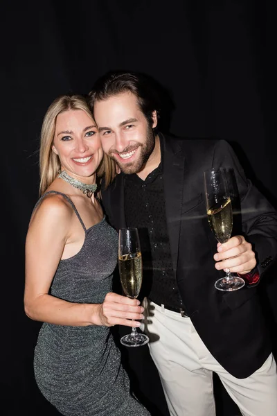 Cheerful man holding glass of champagne near stylish blonde girlfriend isolated on black 