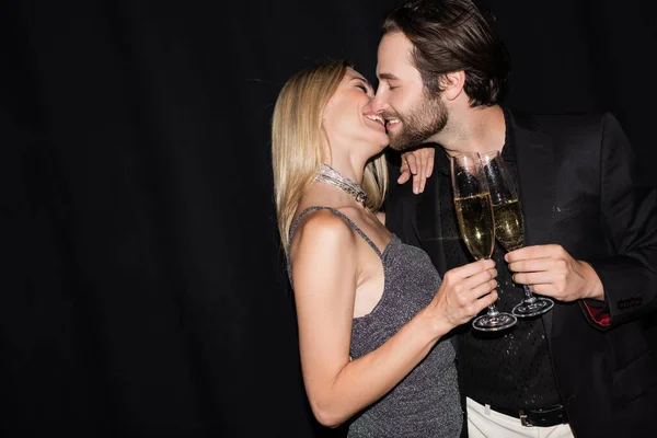 Side view of smiling stylish couple kissing while holding champagne isolated on black
