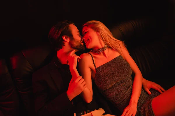 Side view of smiling woman in dress kissing boyfriend on leather couch with lighting isolated on black