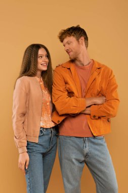 happy teenage girl looking at redhead boyfriend standing with crossed arms isolated on beige 