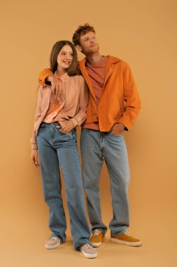 full length of happy young man in autumnal outfit and gumshoes hugging girlfriend on beige