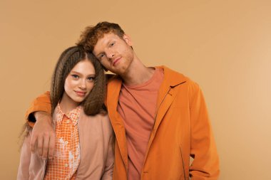 redhead young man in orange jacket hugging cheerful girlfriend isolated on beige