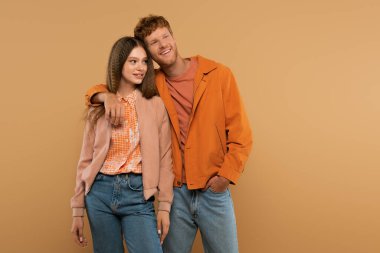 redhead young man standing with hand in pocket and hugging cheerful girlfriend isolated on beige