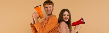 cheerful young couple holding loudspeakers and standing isolated on beige, banner