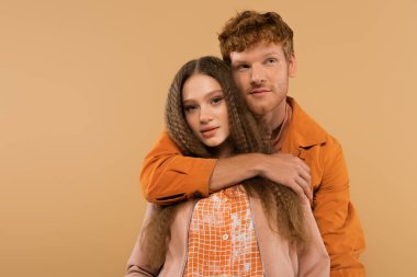 young redhead man hugging girlfriend with wavy hair isolated on beige