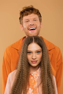 joyful man with red hair smiling behind girlfriend with wavy hair isolated on beige