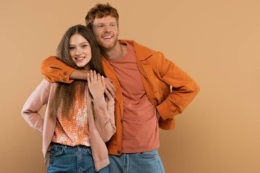 young happy man with red hair looking at camera and hugging pretty girlfriend isolated on beige