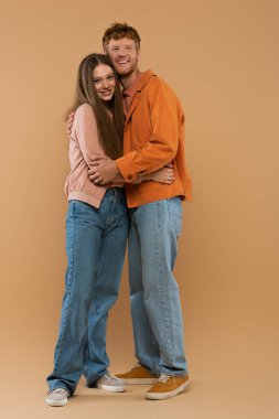 full length of young and cheerful couple hugging while standing on beige