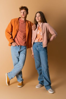 full length of cheerful and young couple in denim jeans standing on beige