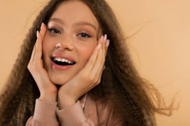 amazed teenage girl looking at camera while touching face isolated on beige