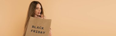 smiling teenage girl holding paper bags with black friday lettering isolated on beige, banner