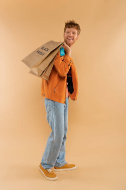 cheerful young man with red hair holding shopping bags with black friday lettering on beige