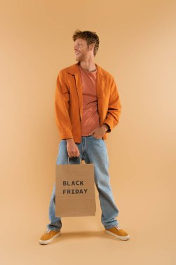 positive young man with red hair holding shopping bag with black friday lettering and posing on beige