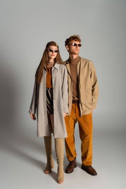 full length of young and stylish couple in autumnal outfits posing on grey