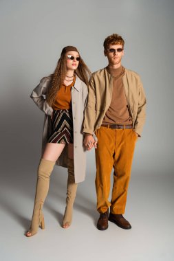 full length of young and stylish couple in autumnal outfits holding hands on grey