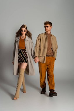full length of young couple in stylish autumnal outfits and sunglasses holding hands while walking on grey