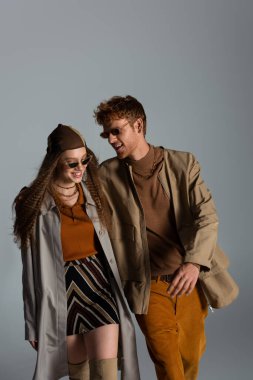happy young couple in autumnal outfits and sunglasses smiling isolated on grey