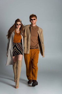 full length of young and stylish couple in autumnal outfits walking together on grey