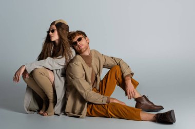 full length of young and stylish couple in autumnal outfits sitting together on grey