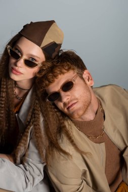 young and stylish couple in sunglasses and autumnal outfits looking at camera isolated on grey