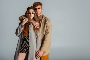 redhead man in sunglasses hugging stylish girlfriend in skirt and trench coat isolated on grey