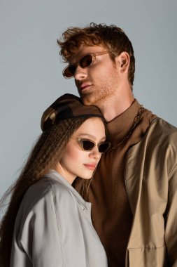 young couple in sunglasses and autumnal clothes standing together isolated on grey