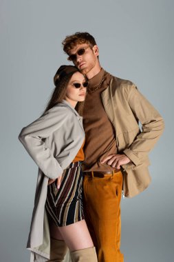 young couple in sunglasses and autumnal clothes posing together isolated on grey