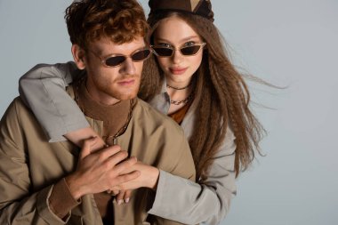 teenage girl in sunglasses hugging stylish and redhead boyfriend isolated on grey