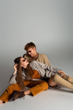 stylish teenage model in sunglasses lying near cheerful young man on grey