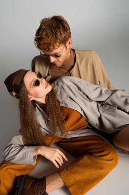redhead young man looking at stylish teenage model in sunglasses lying on grey