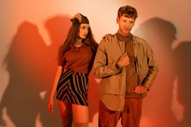 redhead young man posing with stylish model in headscarf and skirt on red background