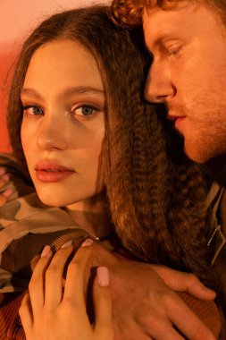 portrait of young man with closed eyes hugging pretty girlfriend looking at camera on red 