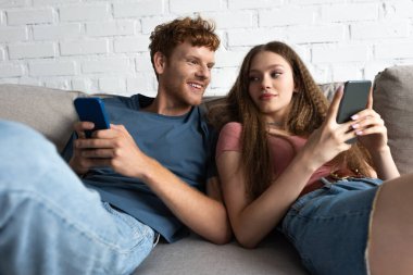 happy young couple using smartphones while sitting on couch in living room